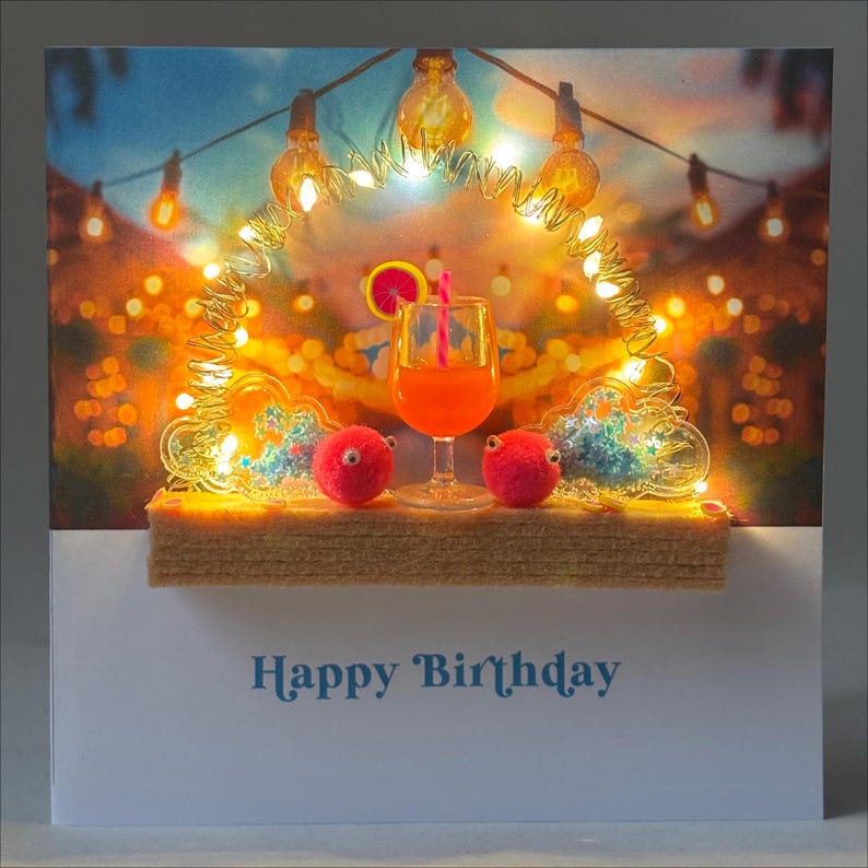May include: A birthday card featuring a cocktail in a glass, two red pom-pom creatures with googly eyes, and string lights. The card has the words "Happy Birthday" in blue script.
