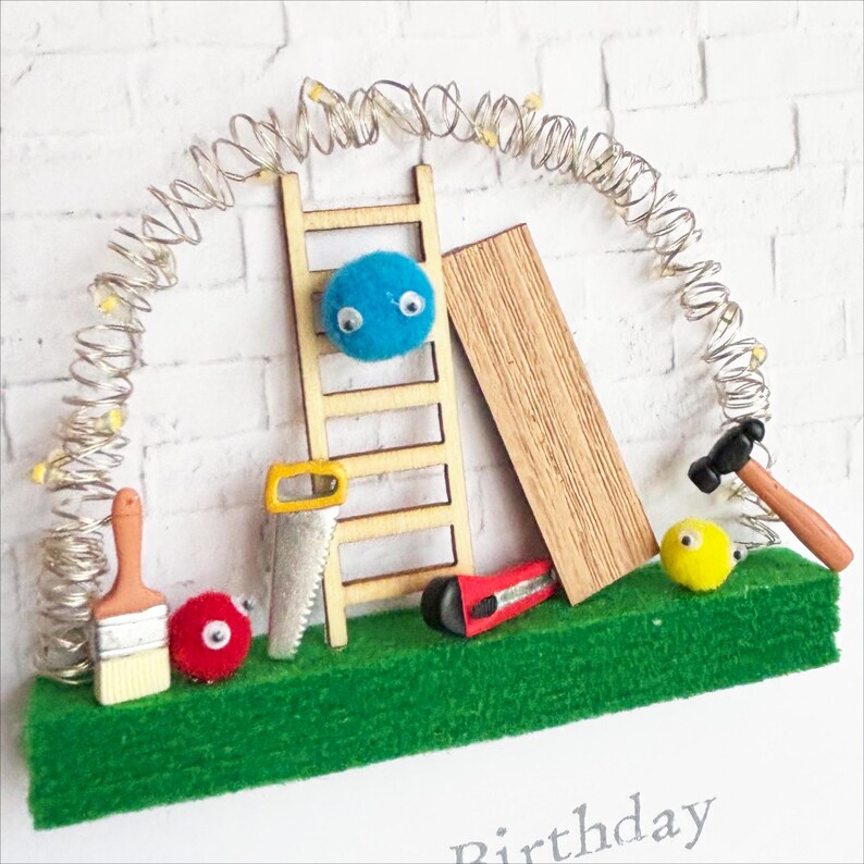 May include: A whimsical construction-themed birthday card. Features a wooden ladder, saw, hammer, and other tools on a green base. A string of lights arches over the scene against a white brick wall. Includes a blue and yellow pom-pom with googly eyes.