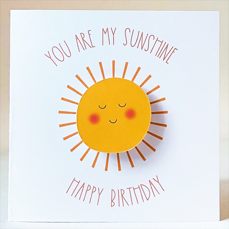 May include: A white card with the text "You are my sunshine" in orange lettering. A yellow sun with a smiling face and closed eyes is in the center of the card. The text "Happy Birthday" is in orange lettering at the bottom of the card.