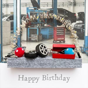 May include: A birthday card with a mechanic theme. The card features a red toolbox, a blue fuzzy ball, a red fuzzy ball, and two toy tyres. The words "Happy Birthday" are printed at the bottom of the card.