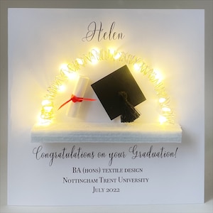 May include: A white graduation card with the name "Helen" in script. It features a miniature mortarboard, diploma, and a string of warm white lights. The text reads "Congratulations on your Graduation!" and details about the degree and university.