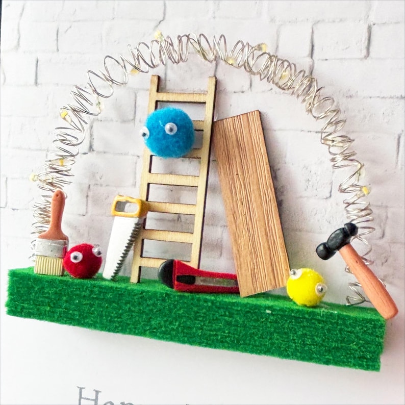 May include: A whimsical diorama featuring miniature carpentry tools and fluffy, googly-eyed creatures. Includes a wooden ladder, saw, hammer, and a strip of wood, all arranged on a green base with a string of lights.
