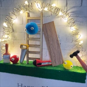 May include: A whimsical diorama featuring miniature tools and felt creatures. A wooden ladder, saw, and hammer are arranged with a blue, red, and yellow felt ball. A string of fairy lights arches over the scene, creating a warm glow.