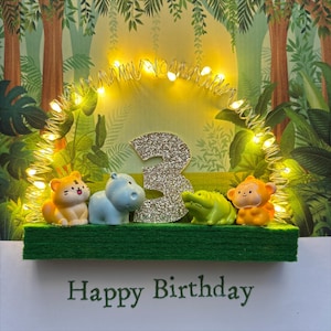 May include: A birthday card with a jungle theme. The card features a green felt base with a tiger, hippo, crocodile, and monkey figurine. A glittery number 3 is in the center, surrounded by fairy lights. The words "Happy Birthday" are printed at the bottom.