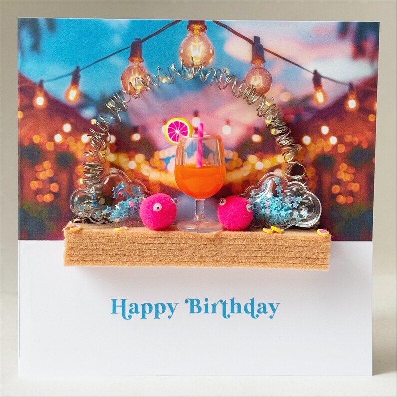 May include: A birthday card featuring a vibrant scene with a cocktail, pink pom-poms, and decorative lights. The card includes the text "Happy Birthday" in a teal font, set against a colorful background with a festive atmosphere.