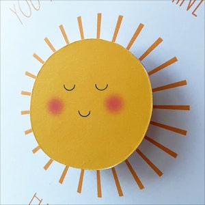 May include: A yellow sun with a smiling face and closed eyes. The sun has a red blush on each cheek. The sun is surrounded by orange rays.