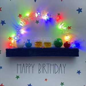 May include: A birthday card featuring a blue felt base and a string of colourful lights forming an arch. There are five small felt figures with googly eyes, two of which are holding glasses of beer. The card reads "Happy Birthday" in black lettering.