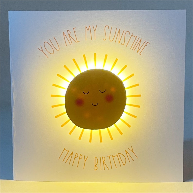 May include: A white card with a yellow sun with a smiling face and closed eyes. The sun is glowing and the card says "You are my sunshine" and "Happy Birthday".