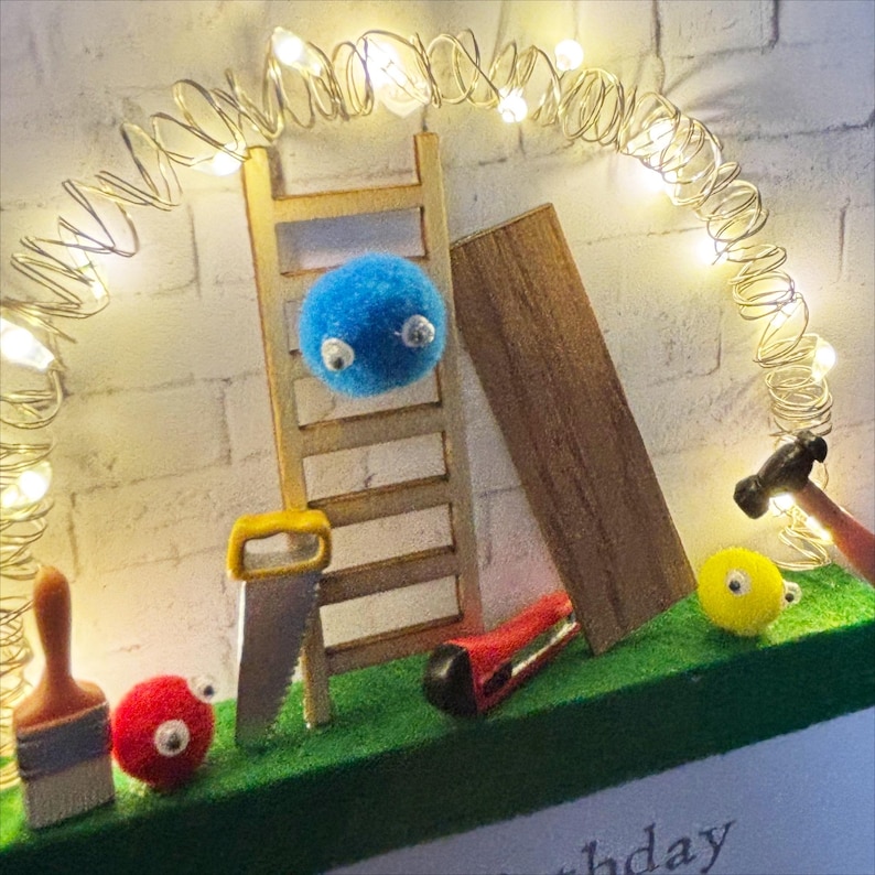 May include: A whimsical diorama featuring miniature tools, a ladder, and fluffy, googly-eyed creatures. A string of fairy lights arches over the scene, with a brick wall backdrop. The scene includes a saw, brush, and hammer.