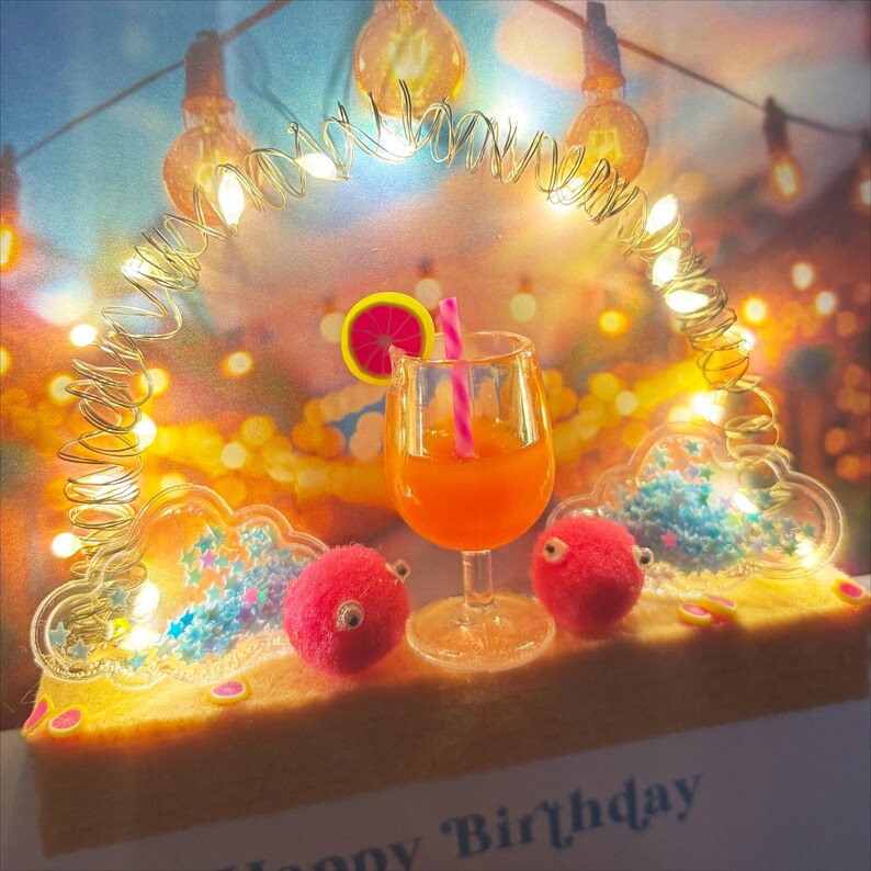 May include: A festive birthday card featuring a cocktail in a glass with a pink straw and a citrus slice. Two pink pom-pom creatures with googly eyes sit on a yellow surface. The card is decorated with fairy lights.