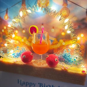 May include: A festive birthday card featuring a cocktail with a pink straw and lemon slice, flanked by two red fuzzy creatures with googly eyes. The card is decorated with fairy lights and the words "Happy Birthday".