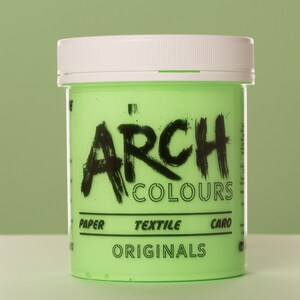 May include: A green plastic jar of paint with a white lid. The jar is labeled "ARCH COLOURS" in black lettering. The jar also says "PAPER, TEXTILE, CARD, ORIGINALS" in black lettering.