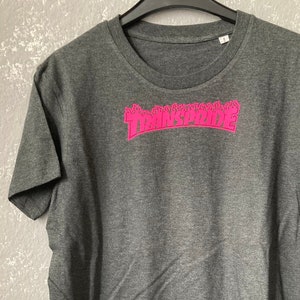 Puede incluir: Una camiseta gris oscuro con un gráfico rosa "TRANSPRIDE" en forma de llama.