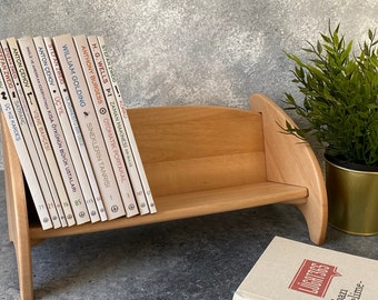 May include: A wooden bookshelf with a light brown finish, holding a collection of books with white spines and black text. The bookshelf is sitting on a gray surface with a small potted plant in the background.