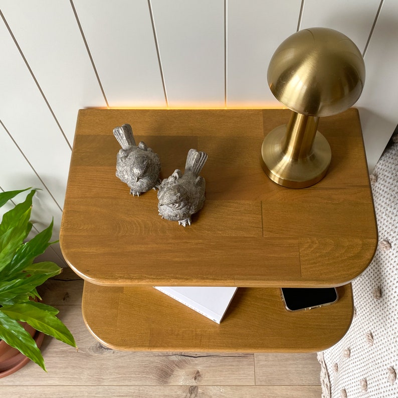 May include: A two-tiered wooden shelf with rounded edges, featuring a gold table lamp with a round shade and two small bird figurines. The shelf is made of light brown wood and has a natural finish.