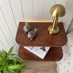 May include: A two-tiered wooden nightstand with rounded edges. The top shelf has a gold lamp shaped like a mushroom, two small bird figurines, and a book titled "Unveiling Light". The bottom shelf has a book titled "The Queen's Gambit".