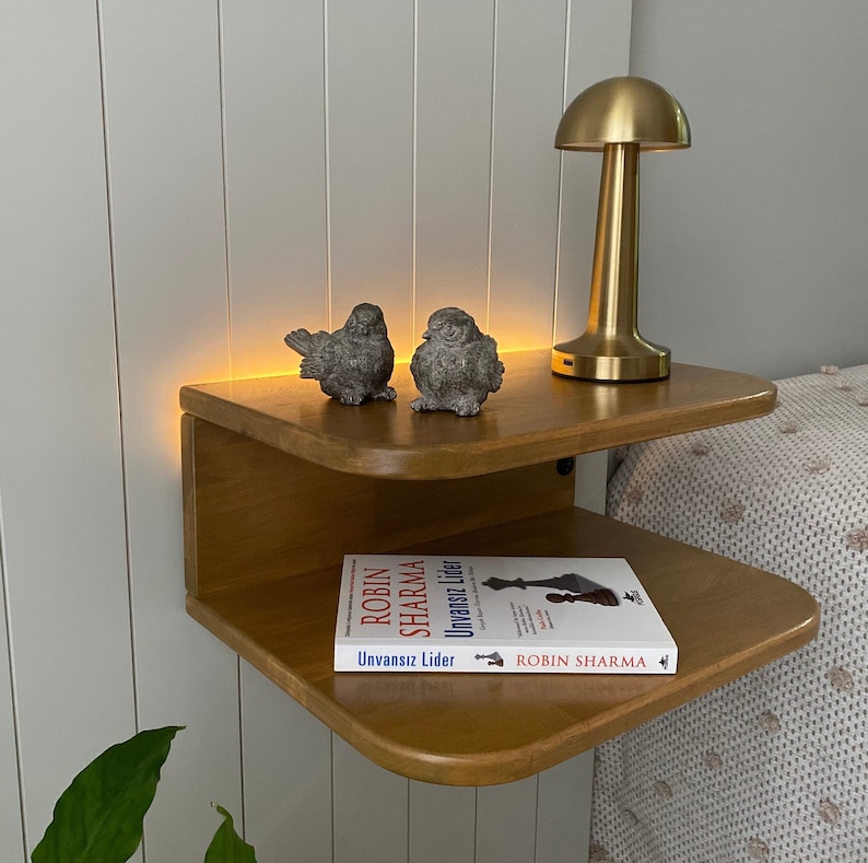 May include: A wooden bedside table with two shelves. The top shelf has a gold mushroom-shaped lamp and two small bird figurines. The bottom shelf has a book titled "Robin Sharma Unvansiz Lider" by Robin Sharma.