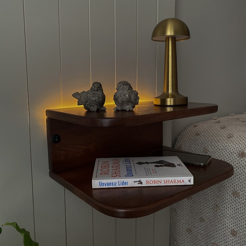 May include: A dark wood floating shelf with two tiers. The top tier has a gold mushroom-shaped lamp and two small bird figurines. The bottom tier has a book titled "Unvansiz Lider" by Robin Sharma. The shelf is mounted on a white wall.