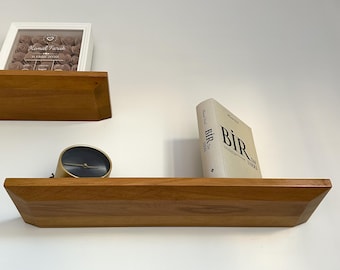 May include: Two wooden shelves mounted on a white wall. The top shelf has a white picture frame with a pink floral design and the text "Kemal Faruk 11.JUNE.2020". The bottom shelf has a gold clock and a book titled "BİR ÇAN SÜRESİ" by "Mehmet Murat Ildan".