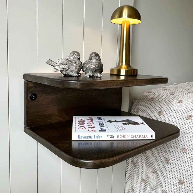 May include: A dark brown wooden floating shelf with two tiers. The top tier has a gold lamp with a mushroom-shaped shade and two small silver bird figurines. The bottom tier has a book titled "Unvansiz Lider" by Robin Sharma.