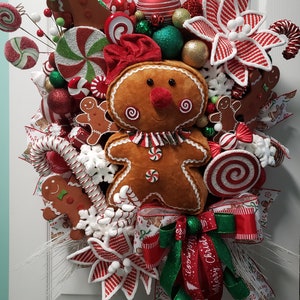 May include: A festive Christmas wreath featuring a plush gingerbread man with a red nose and a striped scarf. The wreath is adorned with candy canes, lollipops, gingerbread men, red and green ornaments, and a decorative bow with "Merry Christmas" text.