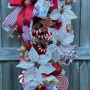 May include: A Christmas swag with a gingerbread man theme. It features a red and white striped ribbon, a red bow, a gingerbread man ornament, and a variety of other gingerbread-themed decorations.