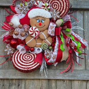 May include: A Christmas wreath featuring a gingerbread person holding a candy cane, surrounded by red and white candy canes, red and white ribbon, and a red bow.