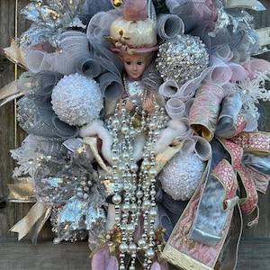 May include: A pink and silver Christmas wreath with a porcelain doll wearing a pink hat and holding a pearl necklace. The wreath is decorated with white and silver mesh, sequins, and a pink velvet bow.