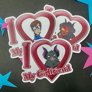 May include: Three die-cut vinyl stickers shaped like red hearts, each featuring the text I Love My Girlfriend in white and red lettering. The stickers display illustrated characters including a person with glasses, a red-skinned demon, and a bat-like creature.