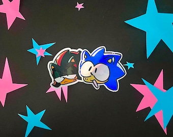 May include: A die-cut sticker featuring a cartoon illustration of the characters Shadow and Sonic the Hedgehog side-by-side. Shadow is black with red accents and a neutral expression, while Sonic is blue with large eyes and a small white mouth.