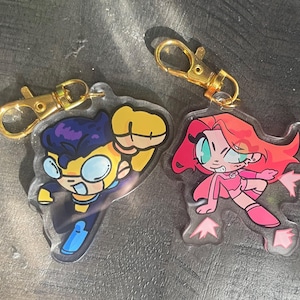 Può includere: Due portachiavi in acrilico con personaggi dei cartoni animati. Un portachiavi raffigura un personaggio in tuta gialla e blu con occhiali, l'altro mostra un personaggio in abito rosa con capelli rossi. Entrambi i portachiavi hanno chiusure color oro.