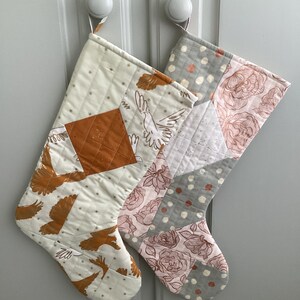 May include: Two quilted Christmas stockings hanging on a white door. The stocking on the left is white with gold stars and a pattern of flying birds. The stocking on the right is grey with white polka dots and a pink floral pattern.
