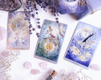 May include: Three tarot cards, The Wheel of Fortune, Knight of Pentacles, and Nine of Cups, are laid out on a white surface with lavender sprigs, pink crystals, and a small glass vial filled with lavender.