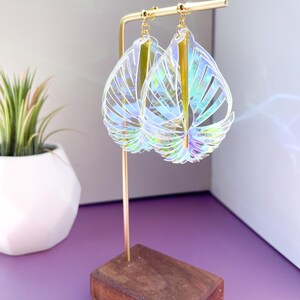 Puede incluir: Un par de pendientes transparentes iridiscentes con forma de hojas y detalles dorados colgando de un soporte de madera.