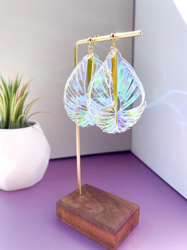 Puede incluir: Un par de pendientes transparentes iridiscentes con forma de hojas y detalles dorados colgando de un soporte de madera.