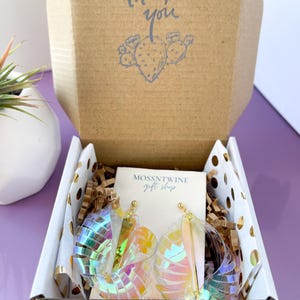 Puede incluir: Una caja blanca con una nota de agradecimiento y una ilustraci&oacute;n de cactus. La caja contiene aretes iridiscentes y una tarjeta peque&ntilde;a que dice "Mossntwine Gift Shop".