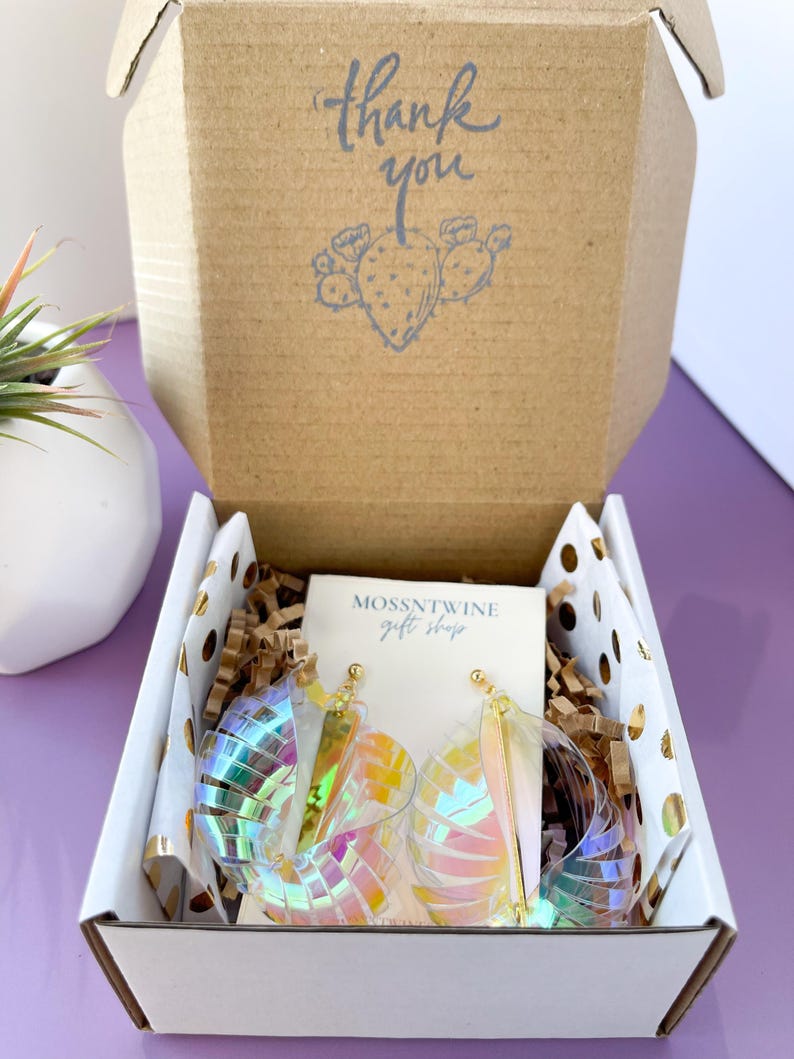Puede incluir: Una caja blanca con una nota de agradecimiento y una ilustraci&oacute;n de cactus. La caja contiene aretes iridiscentes y una tarjeta peque&ntilde;a que dice "Mossntwine Gift Shop".