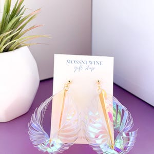 Puede incluir: Un par de pendientes de pl&aacute;stico transparente iridiscente con detalles dorados. Los pendientes tienen forma de alas y cuelgan de una tarjeta blanca con el texto "Mossntwine Gift Shop".
