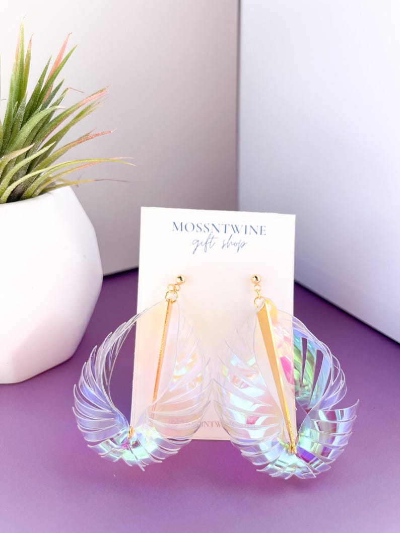 Puede incluir: Un par de pendientes de pl&aacute;stico transparente iridiscente con detalles dorados. Los pendientes tienen forma de alas y cuelgan de una tarjeta blanca con el texto "Mossntwine Gift Shop".