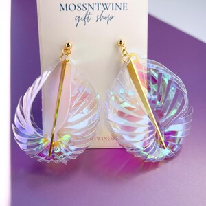 Puede incluir: Un par de pendientes iridiscentes con una barra dorada y un dise&ntilde;o con forma de concha. Los pendientes se muestran en una tarjeta blanca con el texto "MOSSNTWINE gift shop".