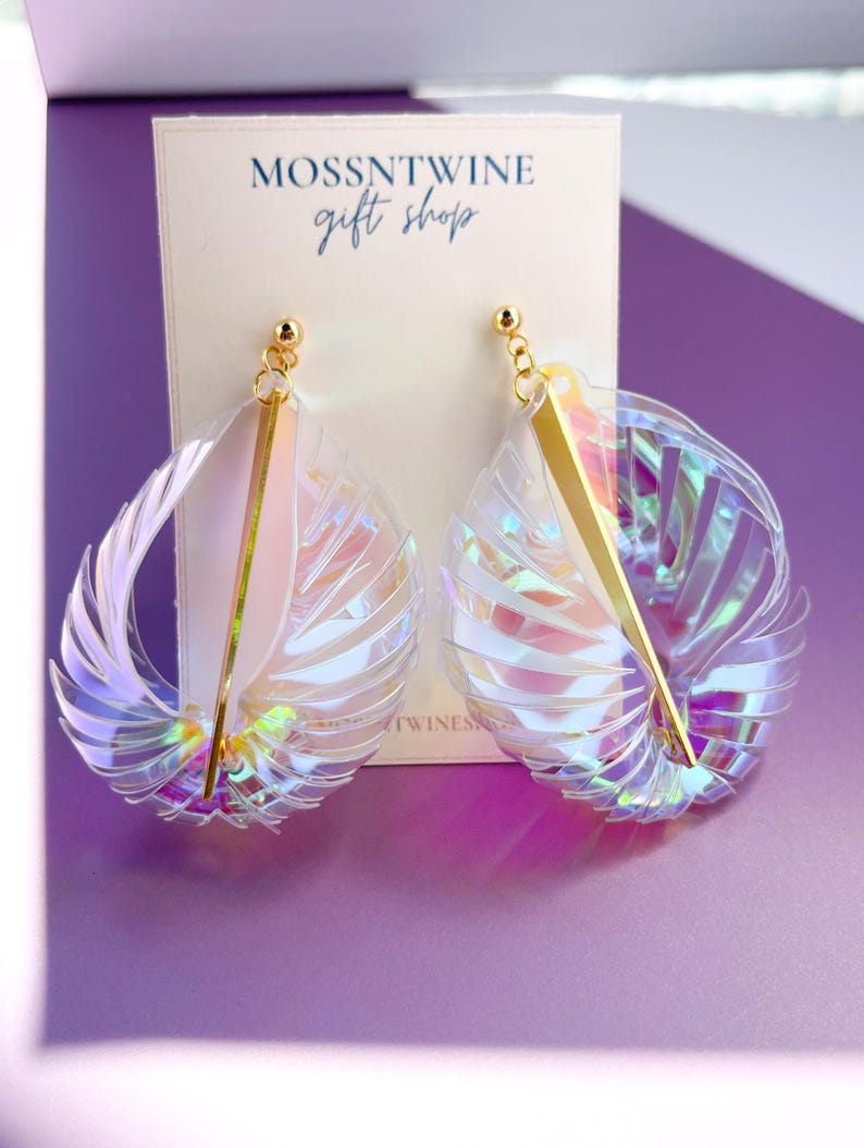 Puede incluir: Un par de pendientes iridiscentes con una barra dorada y un dise&ntilde;o con forma de concha. Los pendientes se muestran en una tarjeta blanca con el texto "MOSSNTWINE gift shop".
