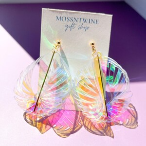 Puede incluir: Un par de pendientes de pl&aacute;stico transparente iridiscente con forma de abanico y una barra dorada en el centro. Los pendientes est&aacute;n sobre una tarjeta blanca con el texto "Mossntwine Gift Shop".
