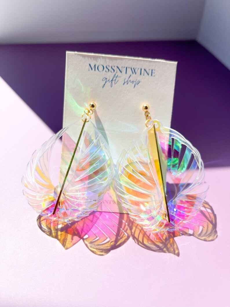 Puede incluir: Un par de pendientes de pl&aacute;stico transparente iridiscente con forma de abanico y una barra dorada en el centro. Los pendientes est&aacute;n sobre una tarjeta blanca con el texto "Mossntwine Gift Shop".