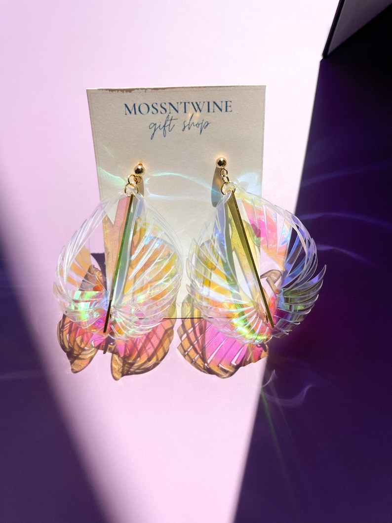 Puede incluir: Un par de pendientes iridiscentes con un acento dorado. Los pendientes tienen forma de abanico y un dise&ntilde;o delicado y femenino. Los pendientes se muestran en una tarjeta blanca con el texto "Mossntwine gift shop".