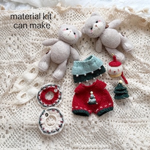 May include: A material kit with two gray knitted cat dolls, along with miniature knitted clothing and accessories. The kit includes a red and green outfit with a Christmas tree, a blue skirt, and a small Christmas tree.