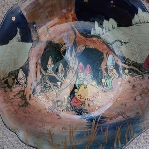 May include: A blue and brown ceramic bowl with a whimsical scene of gnomes under a tree. The gnomes are wearing colorful hats and are sitting on mushrooms. The bowl has a scalloped edge and a gold trim.