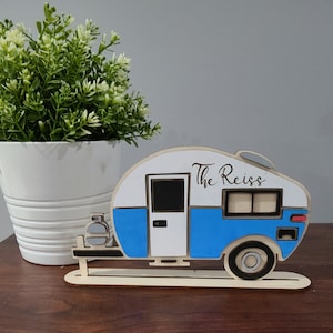 May include: A wooden cutout of a blue and white camper with the text "The Reiss" on the side. The camper is on a wooden base and has a propane tank.