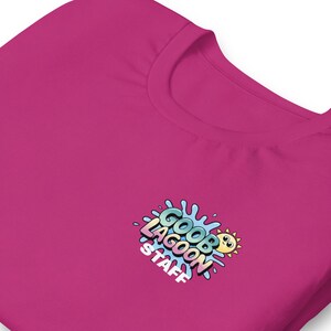 Op de afbeelding: Een magenta crewneck T-shirt met de tekst "GOOB LAGOON STAFF" in een kleurrijk cartoonontwerp. De afbeelding bevat een lachende zon en waterspatselementen. Het shirt is gemaakt van een zacht materiaal.