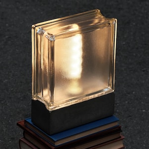 Puede incluir: Una lámpara moderna con una pantalla de bloque de vidrio transparente, iluminada desde adentro, se encuentra sobre una pila de libros antiguos. Los libros tienen cubiertas de varios colores y están colocados sobre una superficie oscura.