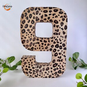 May include: A large number nine, decorated with a leopard print pattern in shades of tan, brown, and black. The number is freestanding and has a hollow center. The background is white, with green foliage accents.